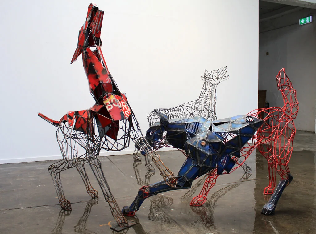 Sculptures of three abstract animals made of wire and recycled materials in red, blue, and metallic shades, titled 'Running with the pack', displayed in a gallery space.