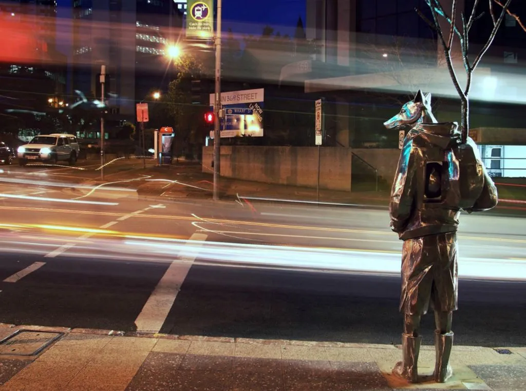 A city intersection during evening with streaks of light from moving vehicles. On the right stands a statue of a figure with a fox head carrying a backpack, under the glow of streetlights, titled 'The Guardian'.