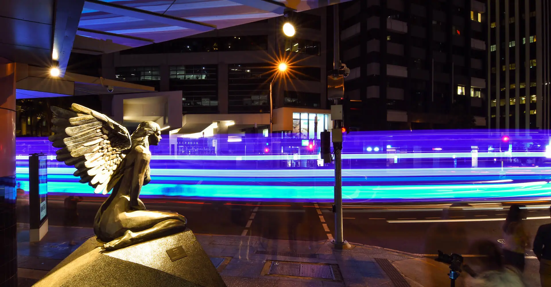 A vibrant cityscape at night with blue and purple streaks of light from moving traffic. In the foreground, a bronze statue of a kneeling figure with large detailed wings, named 'Aurora', is illuminated. Buildings with lit windows and pedestrians blurred in motion fill the background.